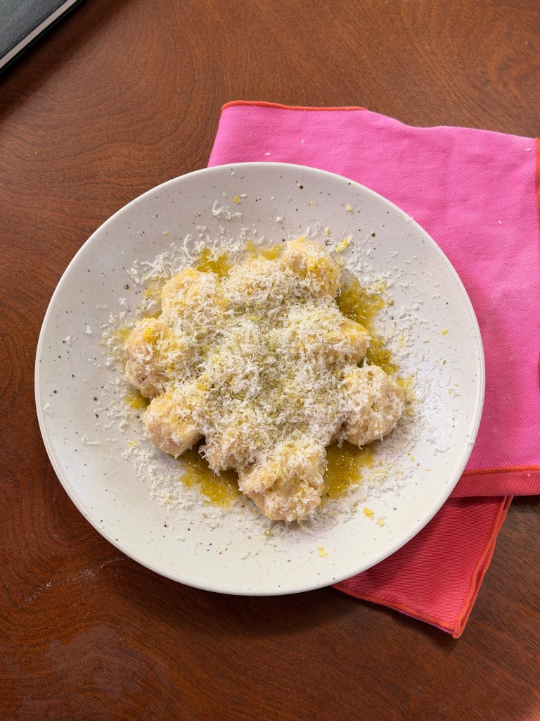 A plate of pasta topped with grated cheese and drizzled with olive oil, placed on a wooden table with a pink cloth underneath.